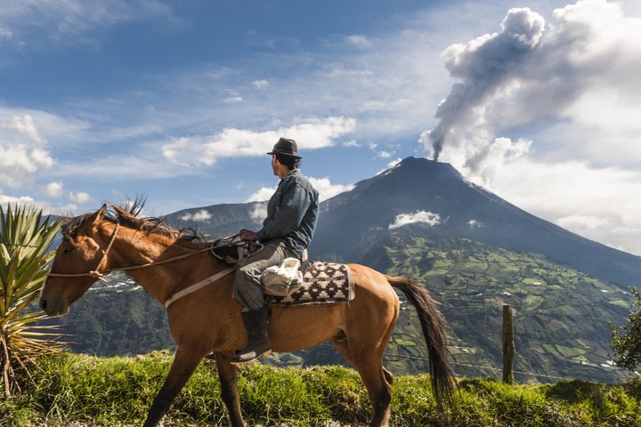 Einheimischer reitet auf seinem Pferd und im Hintergrund raucht es aus dem Vulkan Tungurahua